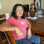 Woman in a pink shirt sits at a wooden dining table, facing the camera, with two others blurred in the background in a home setting.