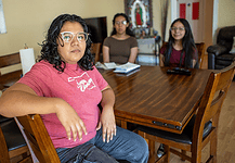Woman in a pink shirt sits at a wooden dining table, facing the camera, with two others blurred in the background in a home setting.