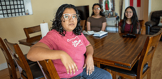 Woman in a pink shirt sits at a wooden dining table, facing the camera, with two others blurred in the background in a home setting.