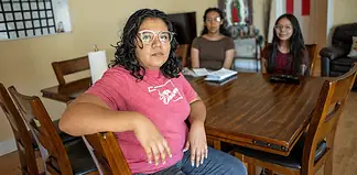 Woman in a pink shirt sits at a wooden dining table, facing the camera, with two others blurred in the background in a home setting.