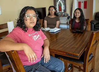 People deported by ICE leave behind families, lasting trauma Woman in a pink shirt sits at a wooden dining table, facing the camera, with two others blurred in the background in a home setting.