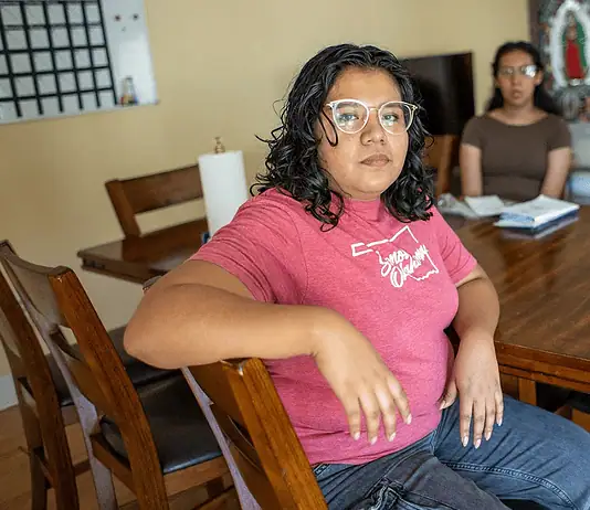 People deported by ICE leave behind families, lasting trauma Woman in a pink shirt sits at a wooden dining table, facing the camera, with two others blurred in the background in a home setting.