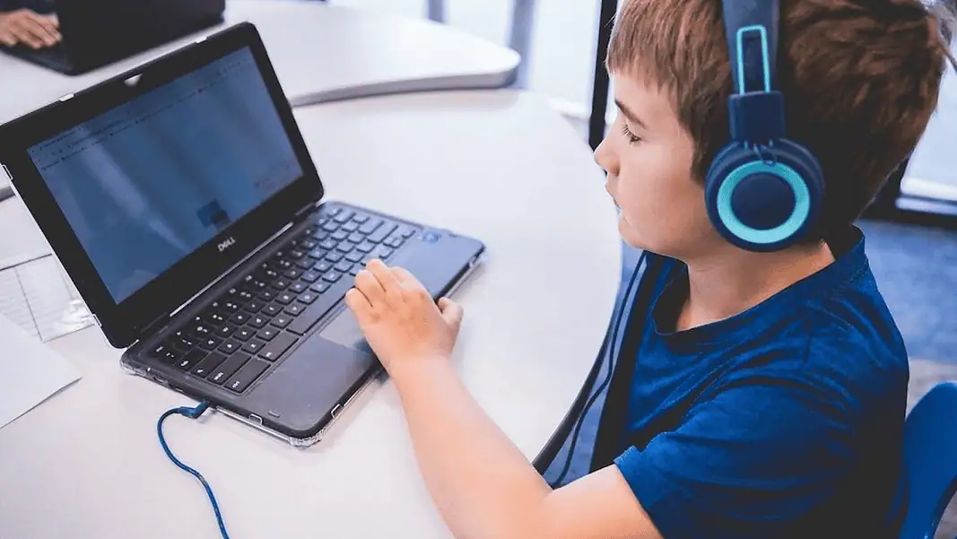 alpha school 1 Child wearing blue headphones typing on a Dell laptop at a white table in a classroom or study area