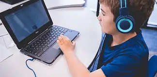 Child wearing blue headphones typing on a Dell laptop at a white table in a classroom or study area