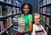 PRESS RELEASE: Summer reading, screen-free fun, and romance authors await in July events at Metro Library Two smiling girls stand in a library aisle, one holding a stack of colorful books for reading and sharing.