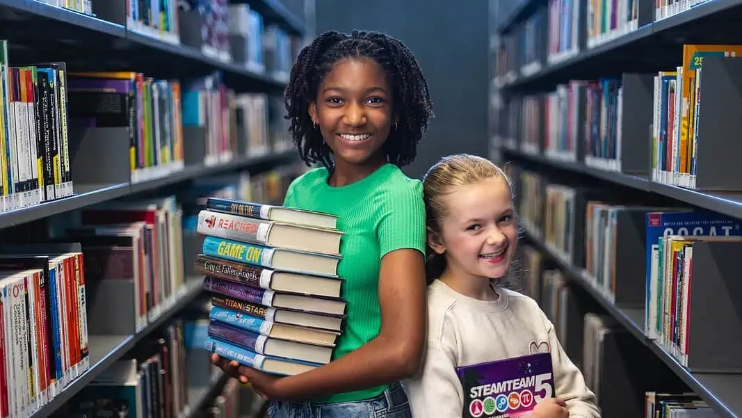 ble_9772 Two smiling girls stand in a library aisle, one holding a stack of colorful books for reading and sharing.