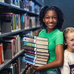PRESS RELEASE: Summer reading, screen-free fun, and romance authors await in July events at Metro Library Two smiling girls stand in a library aisle, one holding a stack of colorful books for reading and sharing.