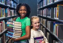 PRESS RELEASE: Summer reading, screen-free fun, and romance authors await in July events at Metro Library Two smiling girls stand in a library aisle, one holding a stack of colorful books for reading and sharing.