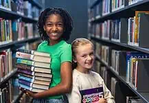 PRESS RELEASE: Summer reading, screen-free fun, and romance authors await in July events at Metro Library Two smiling girls stand in a library aisle, one holding a stack of colorful books for reading and sharing.