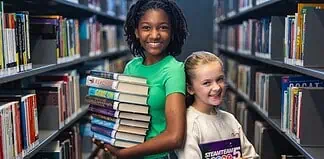 Two smiling girls stand in a library aisle, one holding a stack of colorful books for reading and sharing.