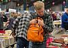 PRESS RELEASE: The Friends of the Library host Summer Book Sale August 14-15 Man in a plaid shirt adjusts an orange backpack while browsing stacks of books at a crowded book sale/fair.