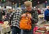 PRESS RELEASE: The Friends of the Library host Summer Book Sale August 14-15 Man in a plaid shirt adjusts an orange backpack while browsing stacks of books at a crowded book sale/fair.
