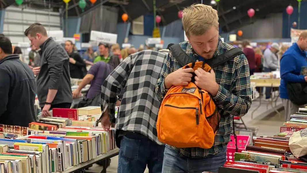 book-sale Man in a plaid shirt adjusts an orange backpack while browsing stacks of books at a crowded book sale/fair.
