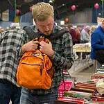 PRESS RELEASE: The Friends of the Library host Summer Book Sale August 14-15 Man in a plaid shirt adjusts an orange backpack while browsing stacks of books at a crowded book sale/fair.