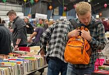 Man in a plaid shirt adjusts an orange backpack while browsing stacks of books at a crowded book sale/fair.