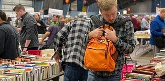 Man in a plaid shirt adjusts an orange backpack while browsing stacks of books at a crowded book sale/fair.