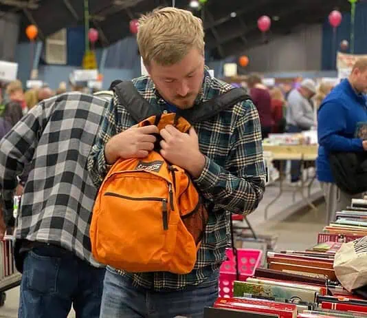 PRESS RELEASE: The Friends of the Library host Summer Book Sale August 14-15 Man in a plaid shirt adjusts an orange backpack while browsing stacks of books at a crowded book sale/fair.