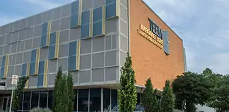Modern brick building with a grey panel facade and blue-yellow decorative panels; a sign on the brick wall reads Transformation Center, with glass windows and trees in front.
