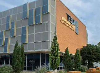 Poor defendants often blocked from OK County pretrial release program Modern brick building with a grey panel facade and blue-yellow decorative panels; a sign on the brick wall reads Transformation Center, with glass windows and trees in front.