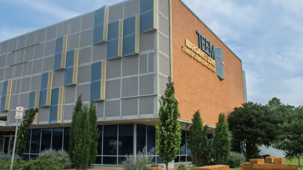 frontier teem 1 Modern brick building with a grey panel facade and blue-yellow decorative panels; a sign on the brick wall reads Transformation Center, with glass windows and trees in front.