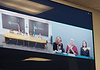 Split-screen video conference of a government meeting: left panel shows officials at a long table with flags in an Oklahoma setting; right panel shows three women, one signing, at a different location.