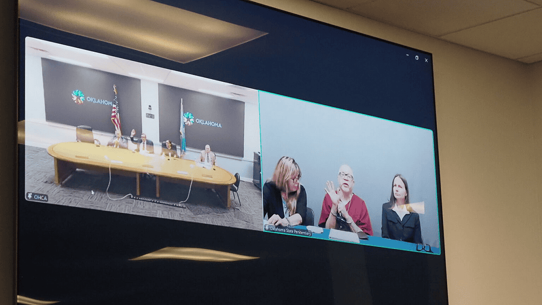Split-screen video conference of a government meeting: left panel shows officials at a long table with flags in an Oklahoma setting; right panel shows three women, one signing, at a different location.