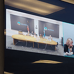 Split-screen video conference of a government meeting: left panel shows officials at a long table with flags in an Oklahoma setting; right panel shows three women, one signing, at a different location.