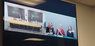 Split-screen video conference of a government meeting: left panel shows officials at a long table with flags in an Oklahoma setting; right panel shows three women, one signing, at a different location.
