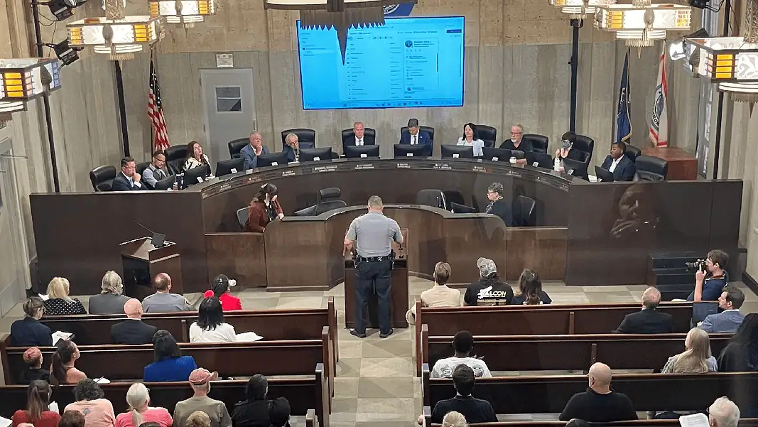 Flock vote Uniformed officer at the podium addressing a city council chamber full of attendees at a curved dais.