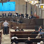 Uniformed officer at the podium addressing a city council chamber full of attendees at a curved dais.