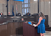 Police confirm threats against City Council following Flock vote Woman in a blue dress speaks at a wooden podium in a city council chamber, while council members sit behind a curved desk with nameplates and flags in the background.