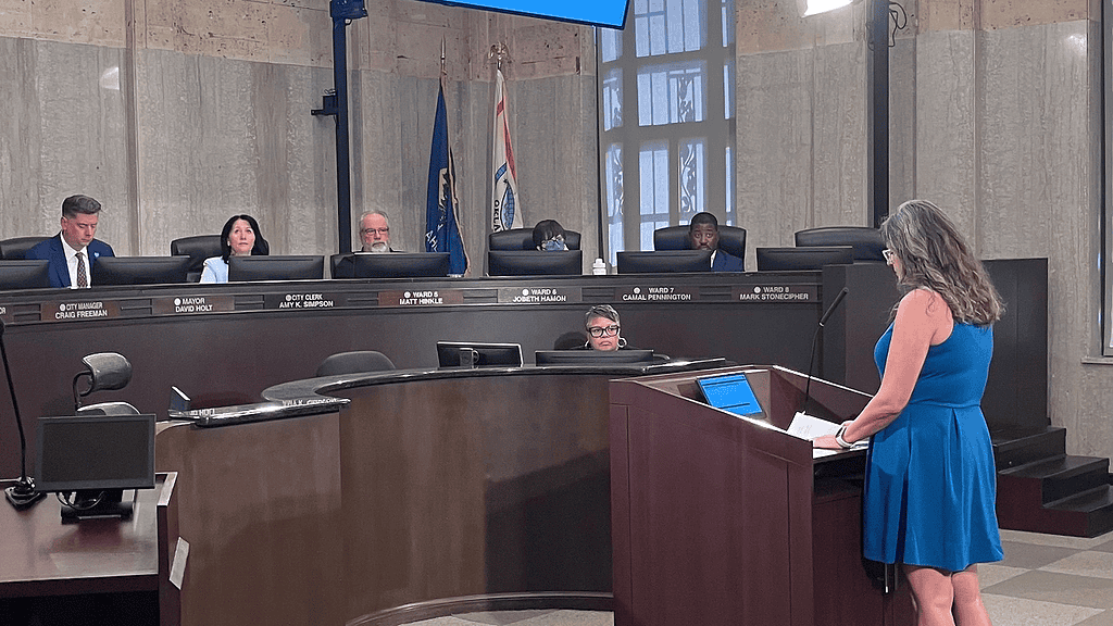 Woman in a blue dress speaks at a wooden podium in a city council chamber, while council members sit behind a curved desk with nameplates and flags in the background.
