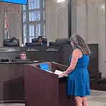 Police confirm threats against City Council following Flock vote Woman in a blue dress speaks at a wooden podium in a city council chamber, while council members sit behind a curved desk with nameplates and flags in the background.