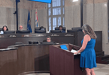 Police confirm threats against City Council following Flock vote Woman in a blue dress speaks at a wooden podium in a city council chamber, while council members sit behind a curved desk with nameplates and flags in the background.