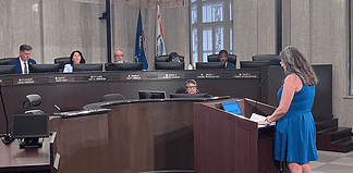 Woman in a blue dress speaks at a wooden podium in a city council chamber, while council members sit behind a curved desk with nameplates and flags in the background.