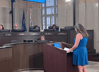 Police confirm threats against City Council following Flock vote Woman in a blue dress speaks at a wooden podium in a city council chamber, while council members sit behind a curved desk with nameplates and flags in the background.