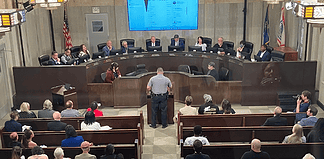 Uniformed officer at the podium addressing a city council chamber full of attendees at a curved dais.