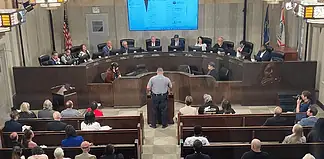 Uniformed officer at the podium addressing a city council chamber full of attendees at a curved dais.