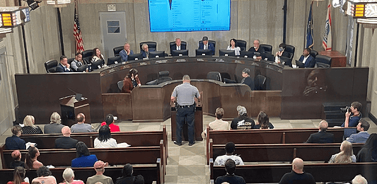 Uniformed officer at the podium addressing a city council chamber full of attendees at a curved dais.