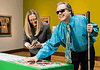 Man with sunglasses and a white cane leaning over a bright green game table, placing a piece as he smiles; woman beside him looks at her phone.