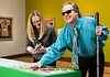 Here’s your guide to accessibility in OKC’s arts institutions Man with sunglasses and a white cane leaning over a bright green game table, placing a piece as he smiles; woman beside him looks at her phone.