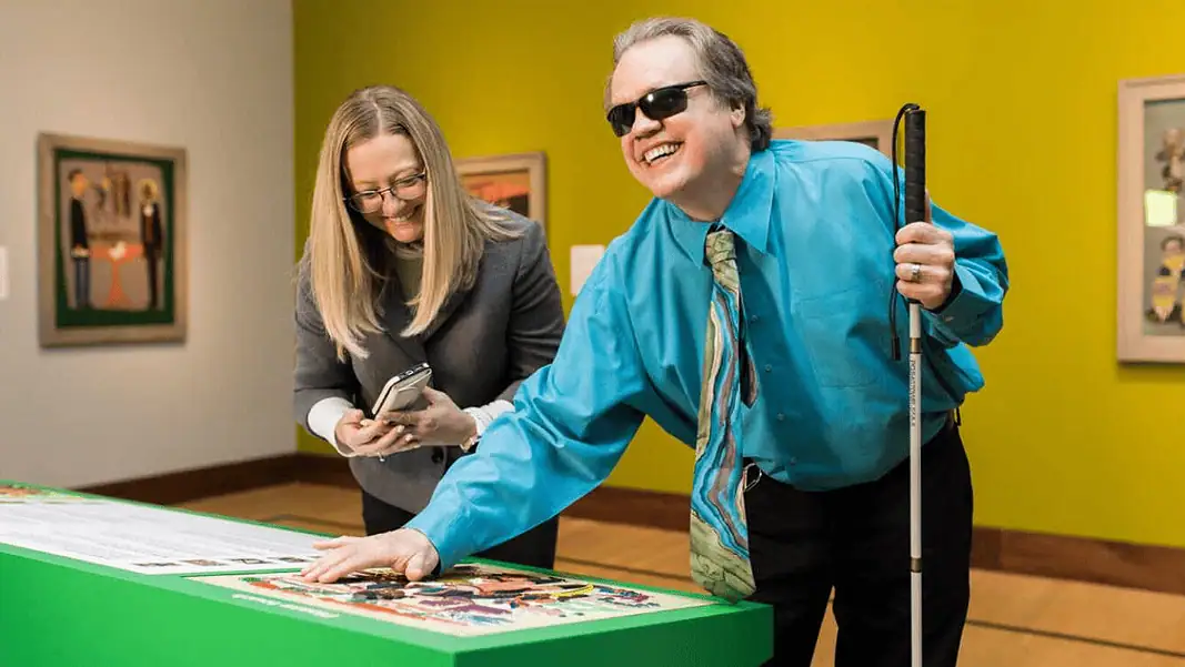 OKCMOA accessibility 2 Man with sunglasses and a white cane leaning over a bright green game table, placing a piece as he smiles; woman beside him looks at her phone.
