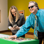 Man with sunglasses and a white cane leaning over a bright green game table, placing a piece as he smiles; woman beside him looks at her phone.