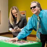 Here’s your guide to accessibility in OKC’s arts institutions Man with sunglasses and a white cane leaning over a bright green game table, placing a piece as he smiles; woman beside him looks at her phone.