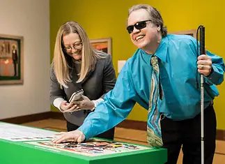 Here’s your guide to accessibility in OKC’s arts institutions Man with sunglasses and a white cane leaning over a bright green game table, placing a piece as he smiles; woman beside him looks at her phone.