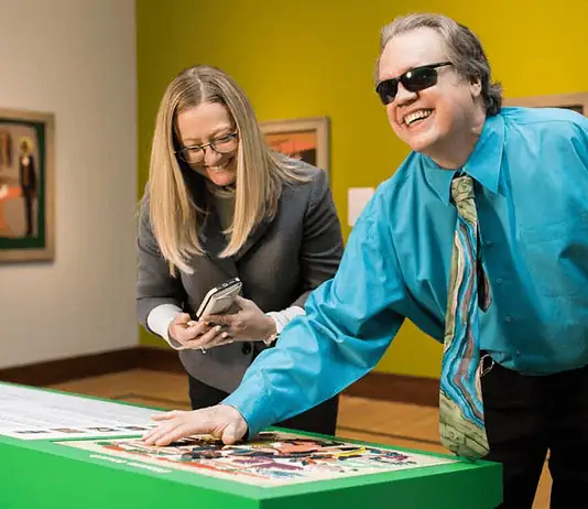 Here’s your guide to accessibility in OKC’s arts institutions Man with sunglasses and a white cane leaning over a bright green game table, placing a piece as he smiles; woman beside him looks at her phone.