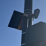 Streetlight pole with a solar panel, security camera, and large control box against a blue sky.