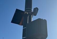 Streetlight pole with a solar panel, security camera, and large control box against a blue sky.