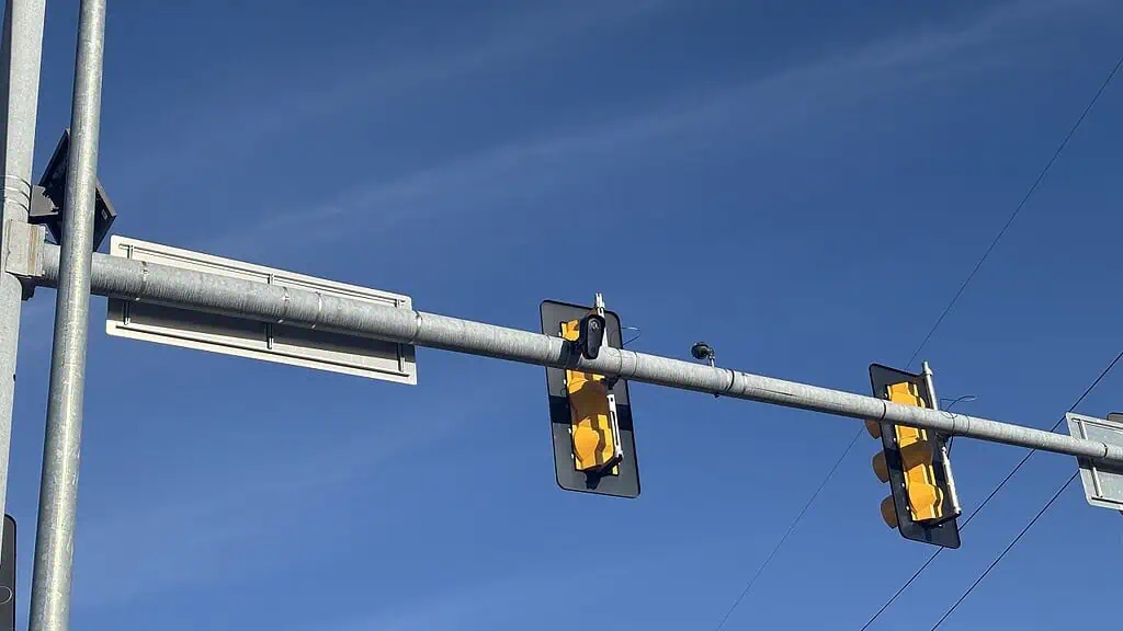Two yellow traffic lights mounted on a horizontal metal bar against a clear blue sky, seen from below over a road.