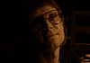 Woman wearing oversized glasses in a dim, sepia-toned room, looking upward with a soft smile under warm lighting.